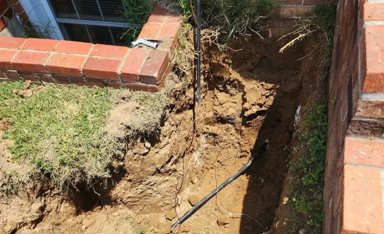 A deep trench dug around a basement window well, exposing pipes and cables for repair or installation.